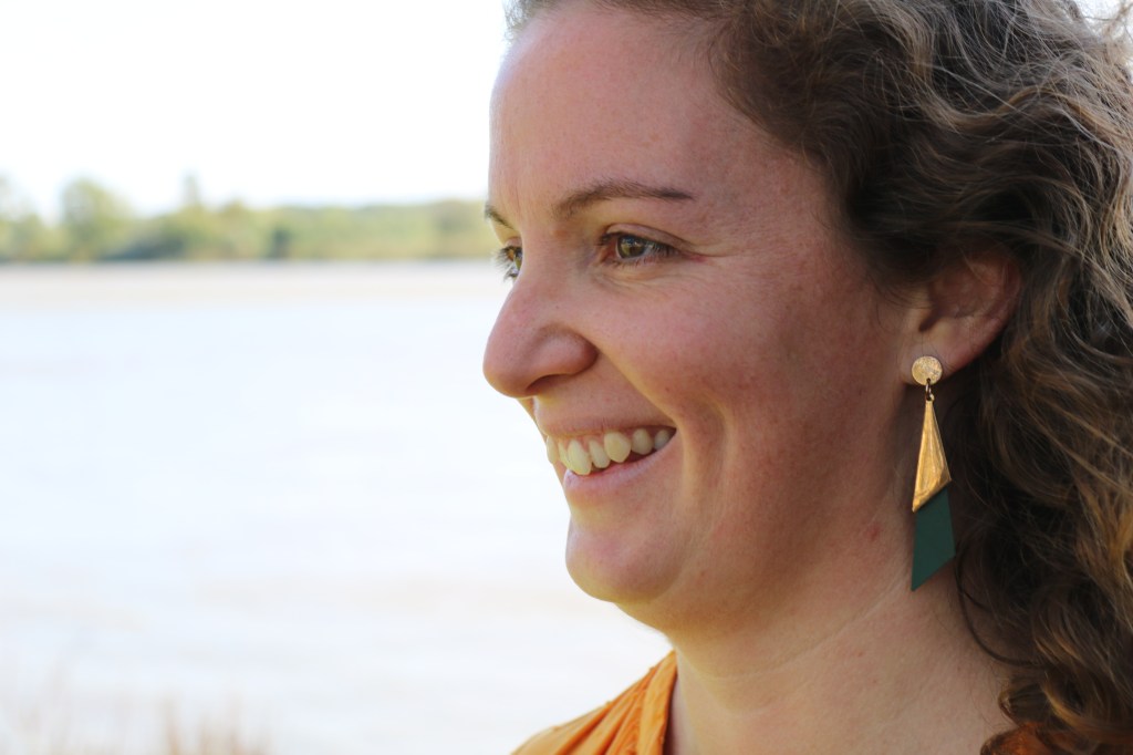 Il s'agit d'une photo d'un visage d'une femme, souriante, de profil. En second plan un distingue une rivière.